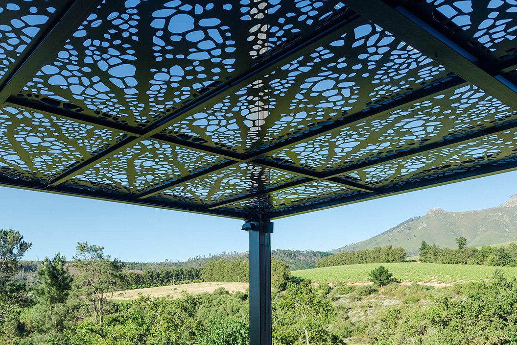 New Pergola at Quoin Rock. Metal structure with crisscross I-beams wrapped with solid wood panels. Laser cut aluminum panels create an organic vine cover shade